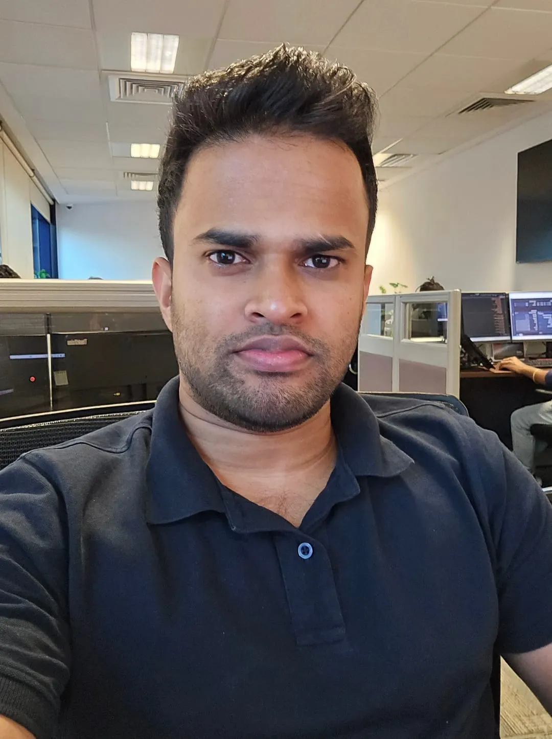 Abhith Rajan in a dark polo shirt, taking a selfie at his desk in an open-plan office